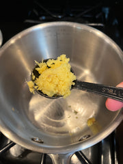 A hand holding a tablespoon of grated ginger over a saucepan