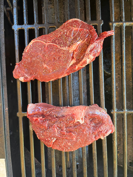 Two raw elk ribeye steaks on the grill