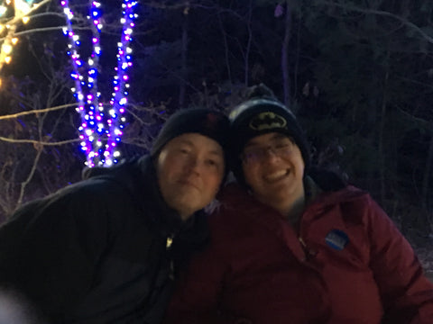 Dan and Amy, a white man and a white woman, wearing glasses, dark hats, and winter jackets, put their heads together for a selfie in front of three trees wrapped in blue and white holiday lights.