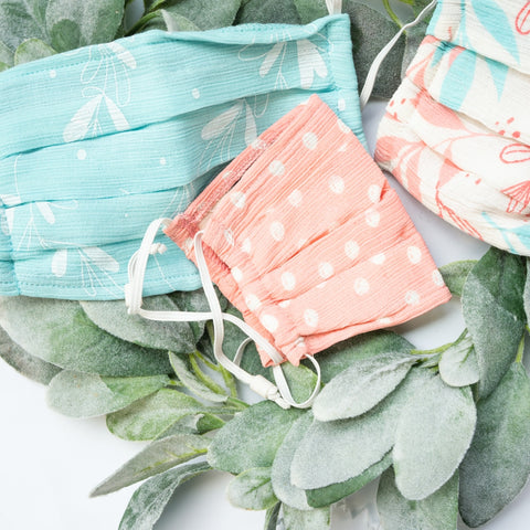 Image of three colorful cloth face masks suggested in the Holiday Gift Guide for Her. All three have different prints. One is light blue with a lead print, another is white with blue and pink leaf print, and the third is pink with white polka dots.