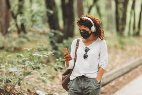 woman walking in trails with headphones on, wearing face mask, looking at phone 