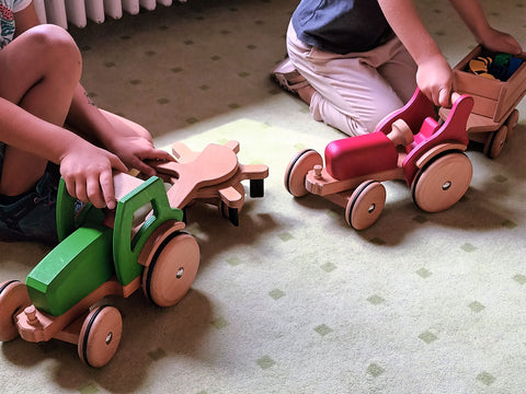 Zwei Kinder spielen mit dem grünen und roten Holztraktor inklusive Anbauteil Heuwender