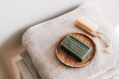 Block of soap on wooden dish next to a wooden nail brush, placed on top of a towel