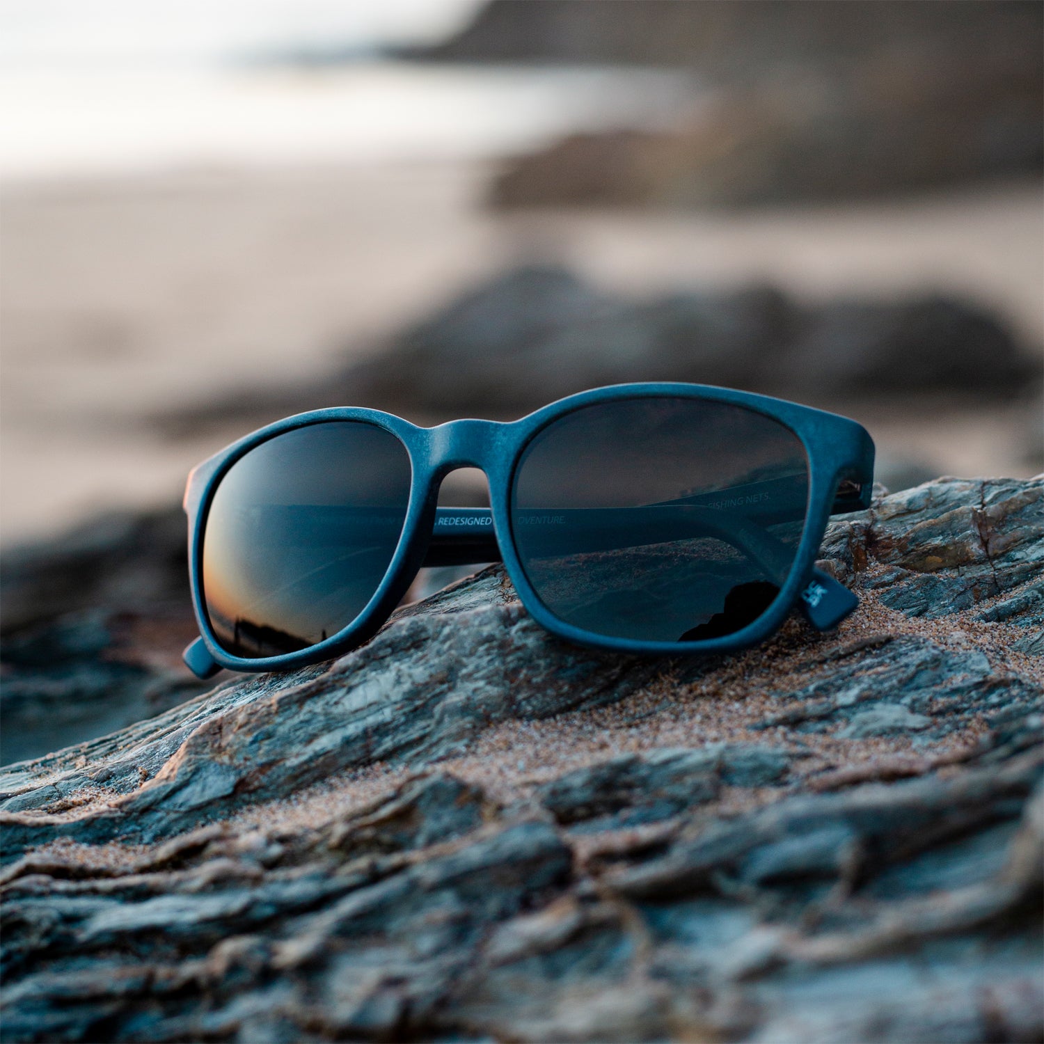 waterhaul fitzroy navy coloured sunglasses on sandy rock with sunset reflected in lenses