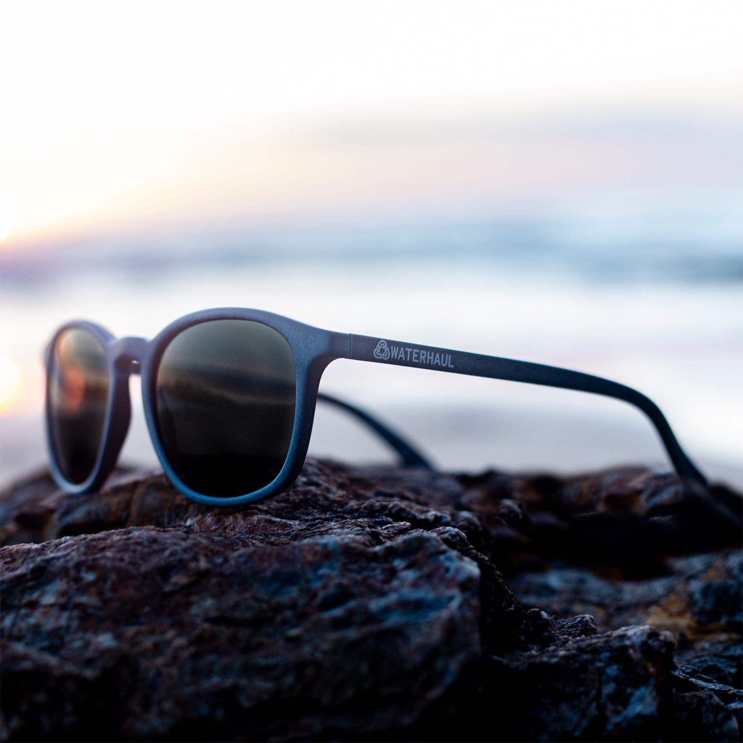 waterhaul kynance navy recycled sunglasses on rocks with sunset reflected in lenses 