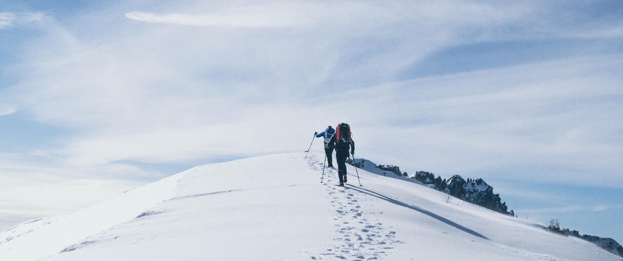 Commerce trends 2023: Photo of two hikers using support poles trekking up a steep, snow-covered slope in the sunshine as they follow the tracks of the hikers who have gone before them.