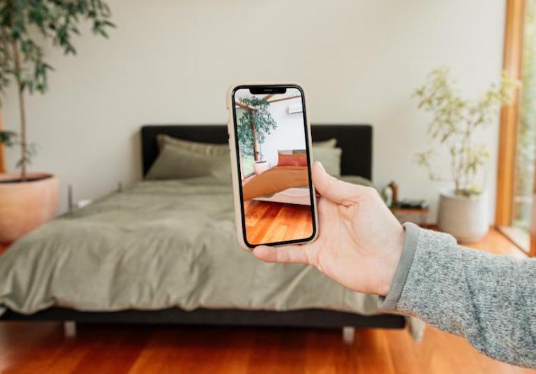 A customer holding a smartphone displaying the AR experience for The Sheet Society, which shows what the selected product would look like on their own bed.