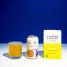 A glass of orange-colored kombucha, a kombucha can labeled "Tangerine & Coffee", and a white bag of "Counter Culture Coffee - Apollo" are placed on a white surface against a blue background.