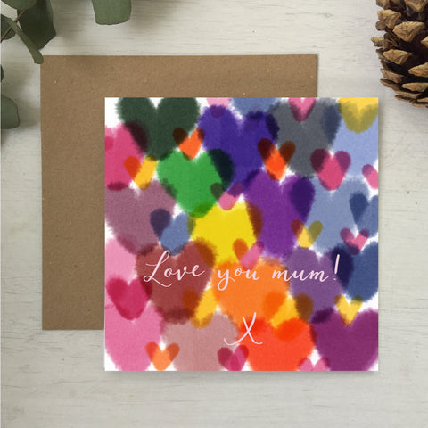 Mother’s Day card covered in multicoloured watercolour hearts and the words ‘love you mum’ are hand lettered in the centre in a light pink colour 