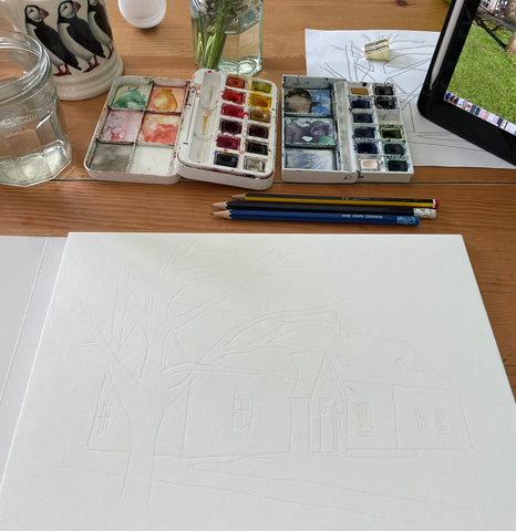 Work desk of a watercolour painter creating a watercolour house portrait - the sheet of paper is surrounded with paints, brushes, a candle, a cup of tea and the iPad showing the photo to paint from