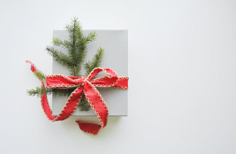 and hope designs christmas gift guide - image of a small white box wrapped in red ribbon with a sprig of evergreen tree on a white background.