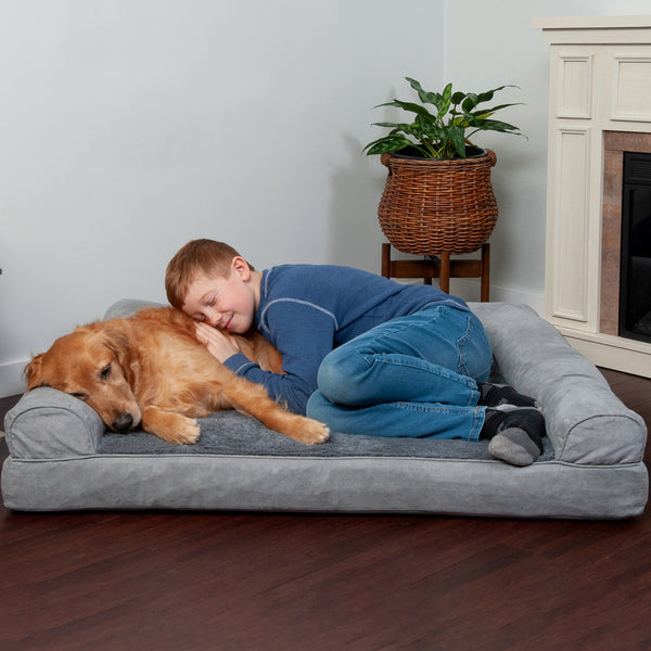 A child in a blue shirt and jeans lying next to a golden colored dog on a FurHaven Pet Bed