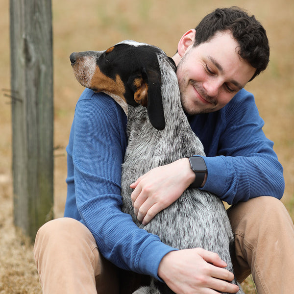 A human in a blue shirt giving a dog with a gray coat and black/brown/white head a hug, at FurHaven Pet Products