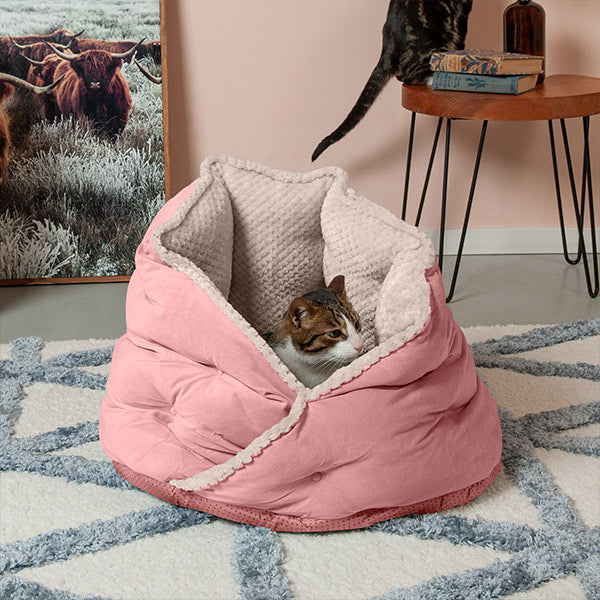 In a pastel room, a brown and white cat sits inside a pink FurHaven Faux Fur & Velvet Hug Pet Bed on the floor.