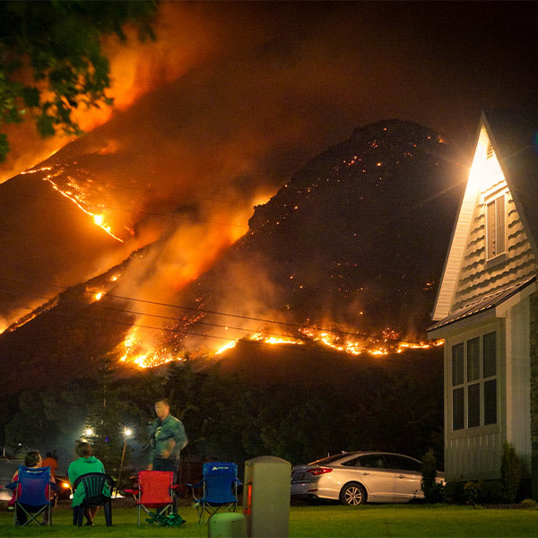 A wildfire rages on a hill while several people sit in camp chairs near their house and observe the phenomenon, at FurHaven Pet Products