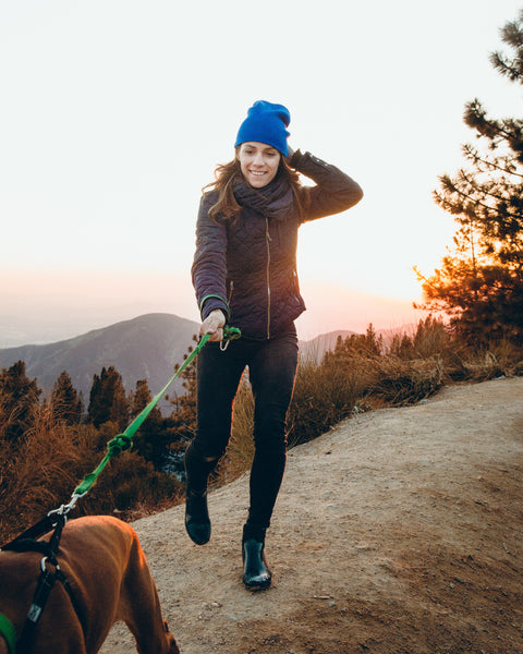 A person wearing a purple jacket and blue beanie walking their brown dog at sunrise in the wilderness, at FurHaven Pet Products