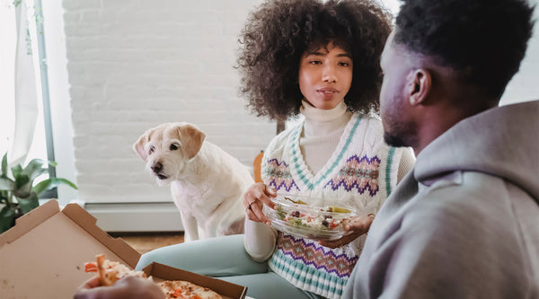 Two people having a meal of salad and pizza, while their blonde dog sits behind them as they chat, at FurHaven Pet Products