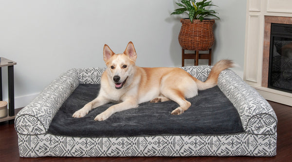 A yellow and white dog lying on top of a gray, patterned FurHaven Southwest Kilim Bed, on top of brown hardwood flooring.