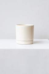 A small glossy white ceramic planter sits on a white surface in a white room. The planter has a matching drainage tray. The planter is empty. It is photographed straight on.
