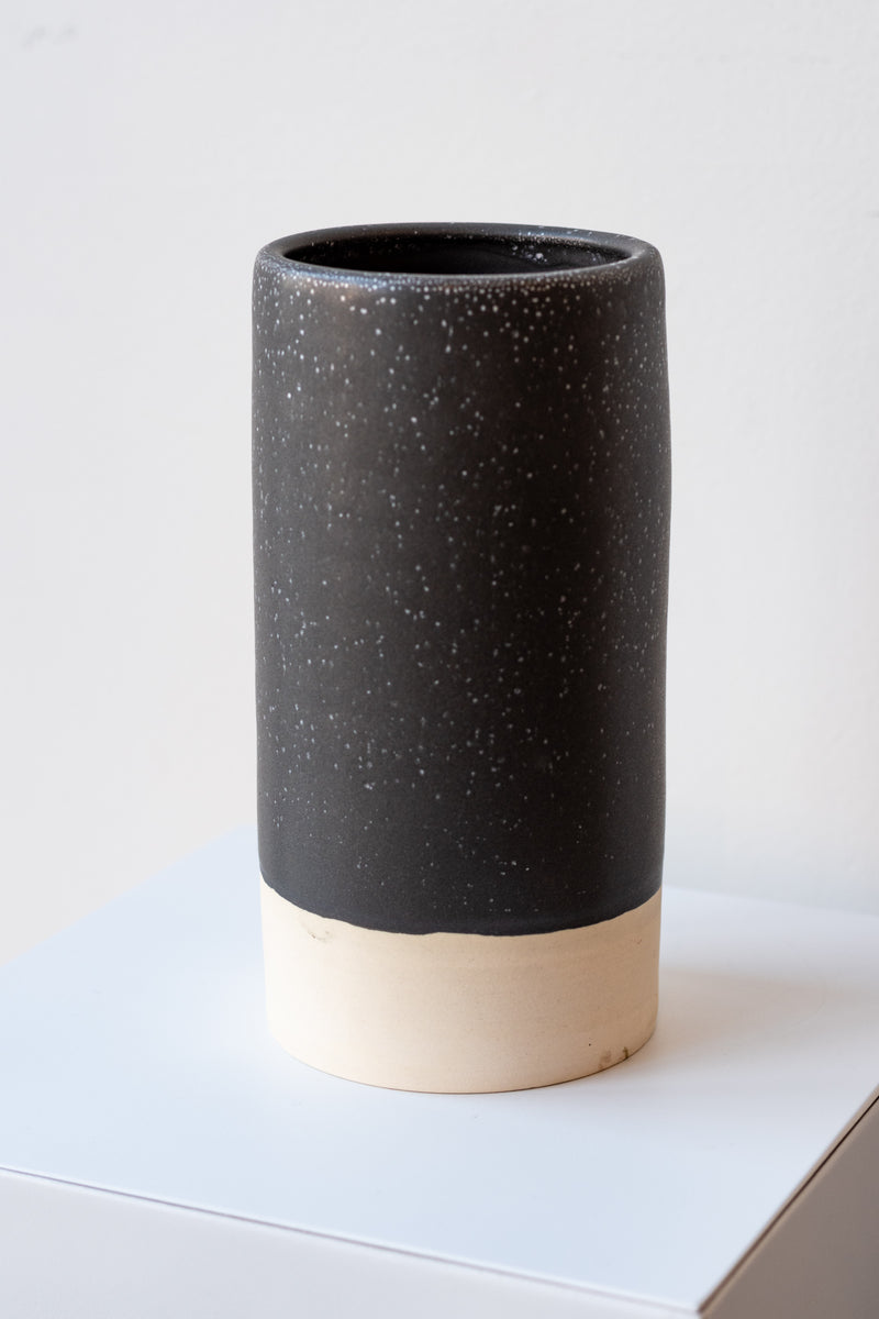 One cylindrical clay vase sits on a white surface in a white room. The vase is glazed with a black glaze with white speckles. The bottom quarter of the vase is unglazed, showing cream-colored clay. The vase is empty. It is photographed closer and at a slight angle. 