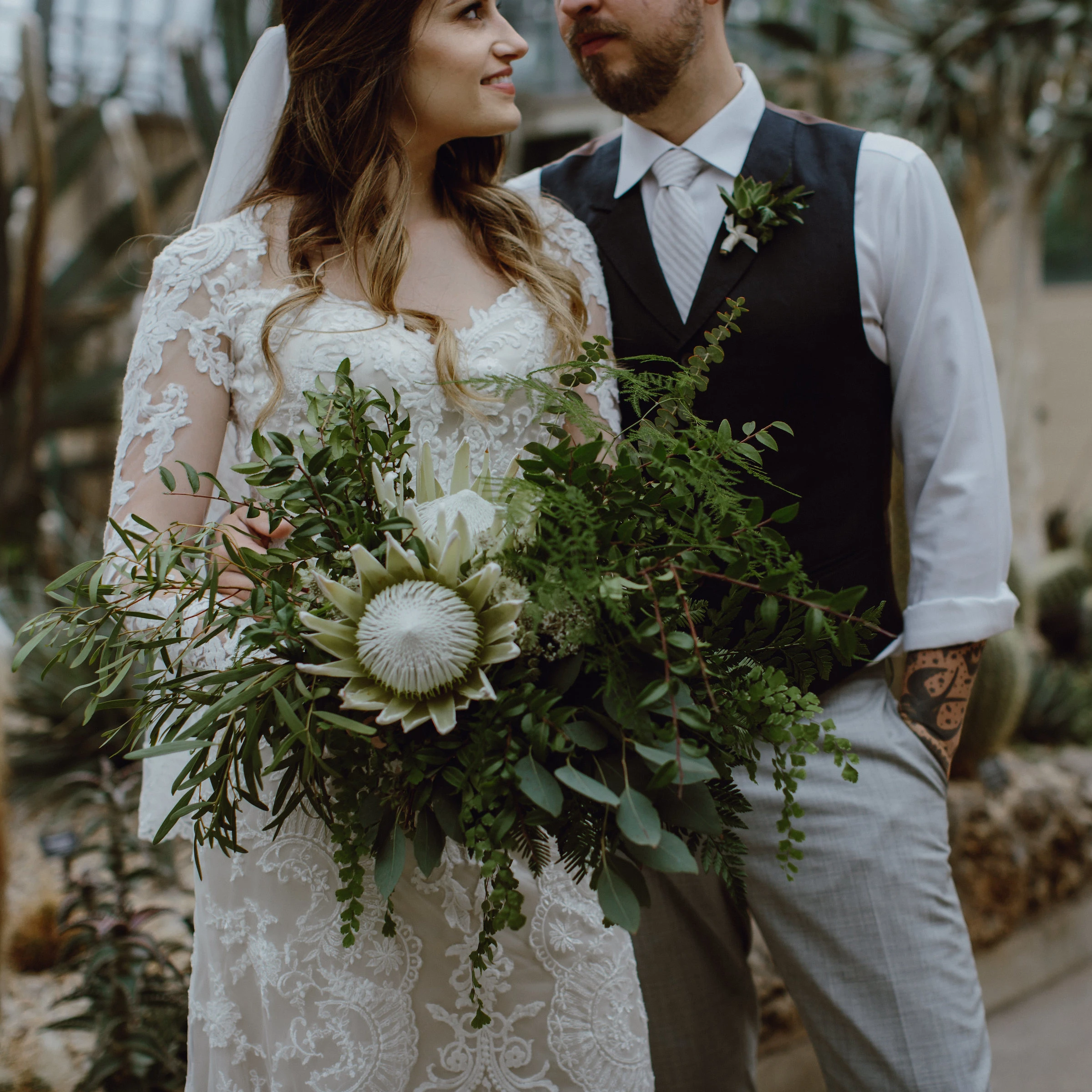 Bride and groom in moody photography style looking at each other. Bride is holding a large greenery arrangement with a large white protea as the focal point