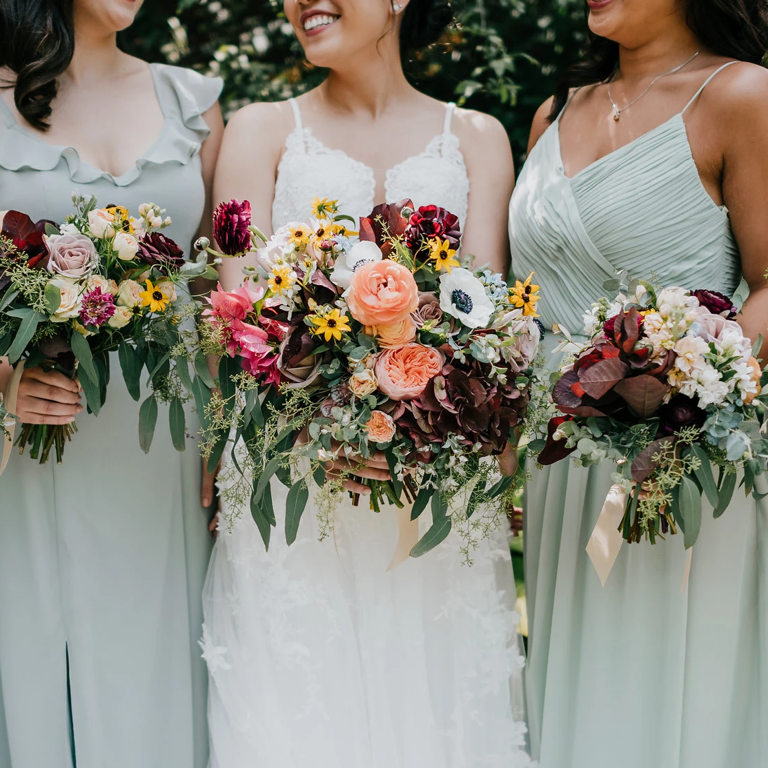 Bride and bridesmaids standing next to each other, each holding a burgundy, pink, white, and yellow bouquet