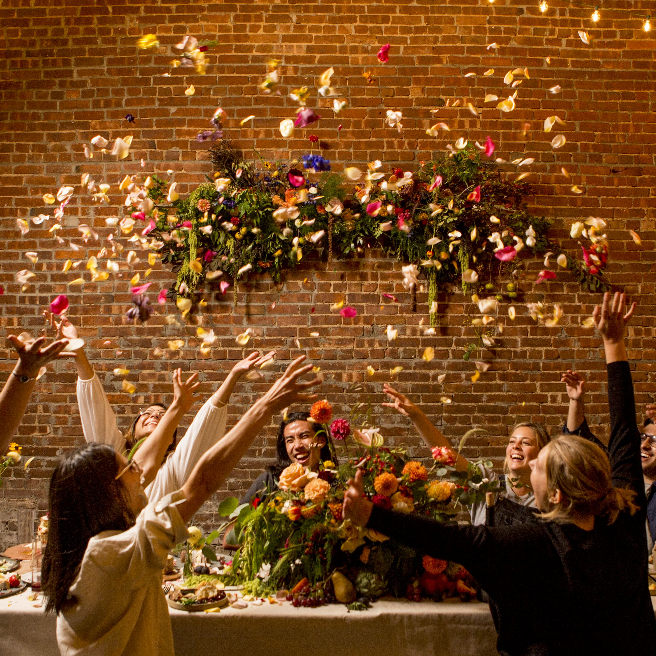 Several fiends sit around a table throwing flower petals in the air. The table has a lush floral centerpiece