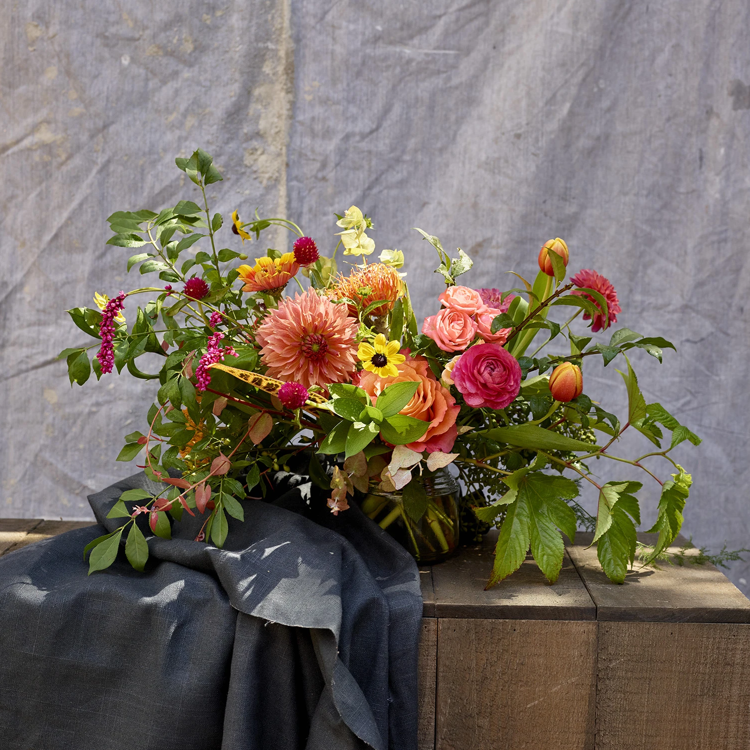 Orange, pink, and yellow floral arrangement sits on a wood surface in front of a grey cloth backdrop