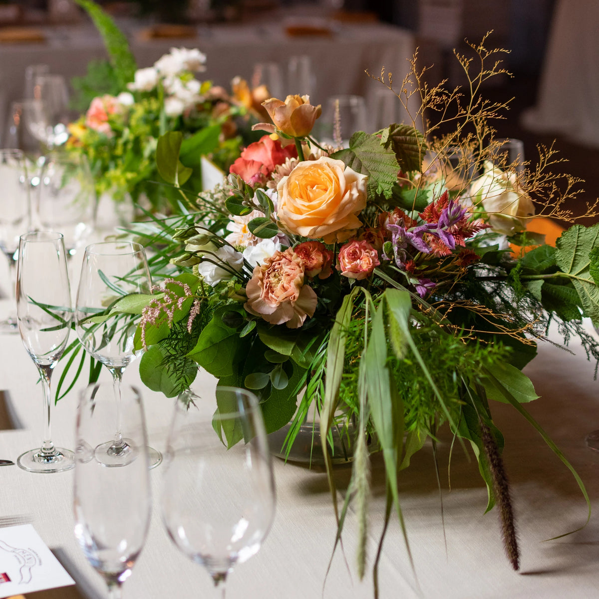 Wedding floral arrangement sits on a white tablecloth surrounded by polished wine glasses