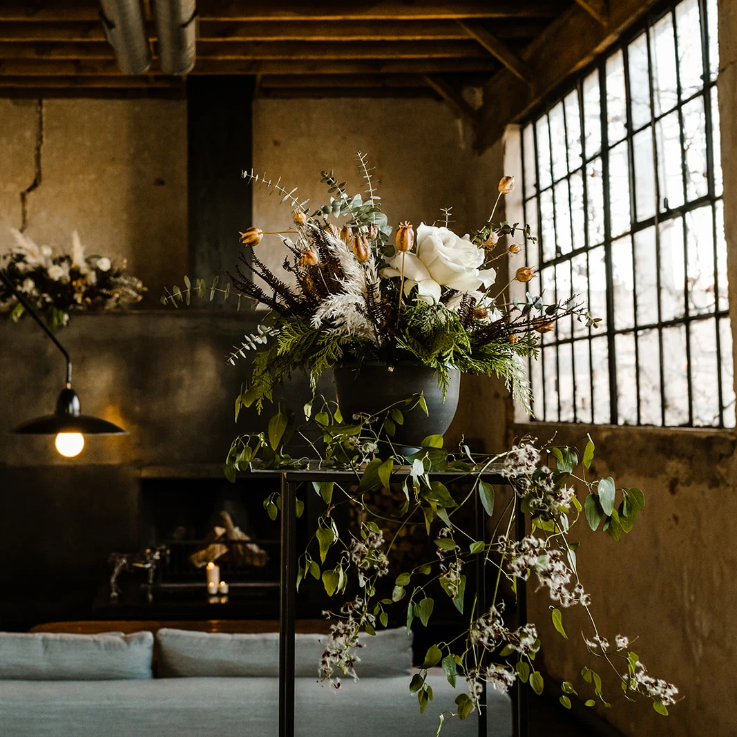 Muted white and beige floral arrangement in a black vessel sits in an industrial-looking room