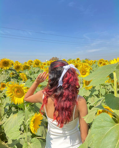 A girl with red hair and a belt accessory which ties her hair whilst she stands in the sunflower field