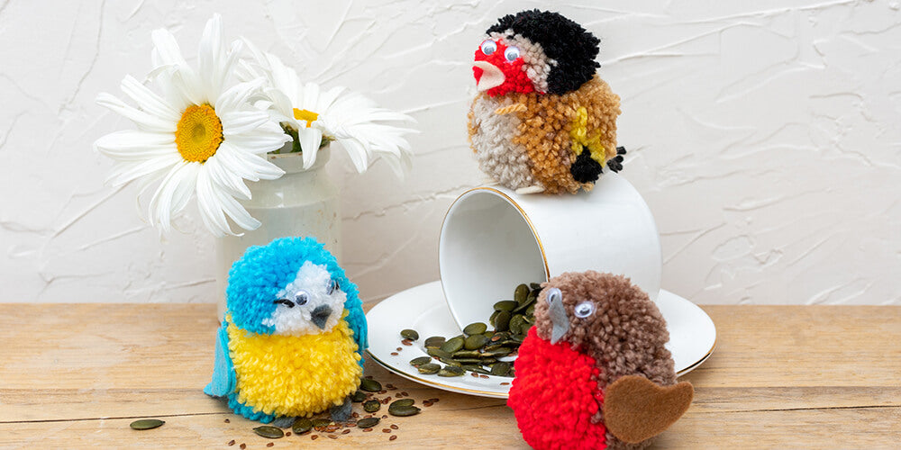 Pom pom garden friends craft kit - a photo of pom pom birds (robin, blue tit and gold finch) on table with bird seeds, upturned mug and flowers in vase.