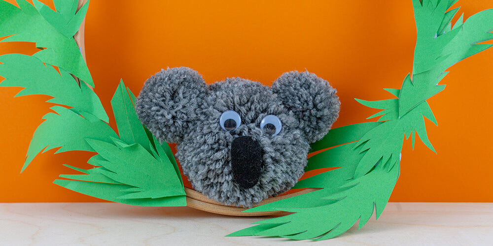 Pom Pom Koala in foliage ring on orange background. Make a pom pom koala with Pom Stitch Tassel's pom pom koala craft kit.