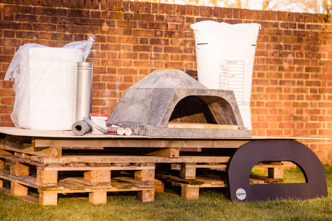All parts of a DIY pizza oven kit placed ontop of pallets in front of a brick wall