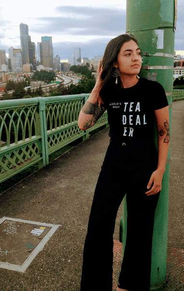 Girl wearing World's Best Tea Dealer Shirt on a bridge with city in background