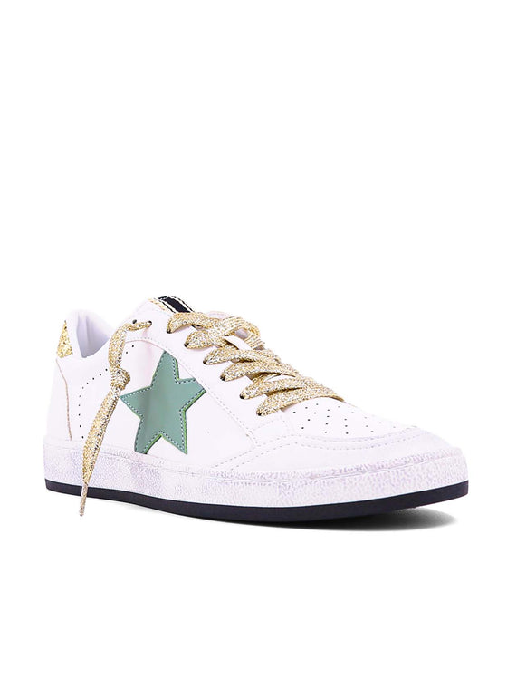 Front/side view of the Paz Sneaker features a white leather upper with a gold lace-up closure. This sneaker is finished with a green star detail, a gold glitter heel detail, and a distressed rubber outsole with a black sole. 
