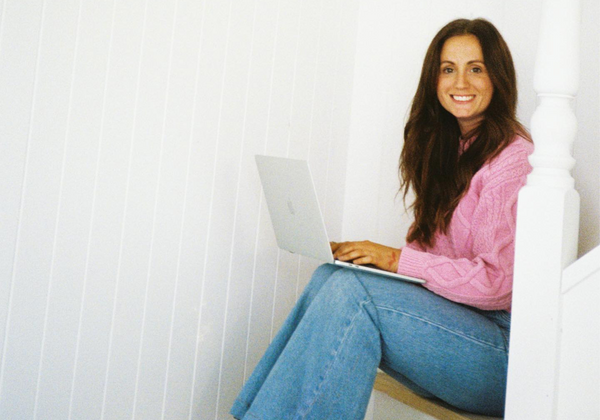 Sam wearing blue jeans and pink top, sitting on white stairs with laptop on knees
