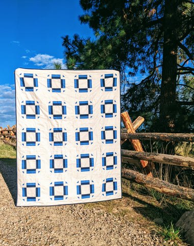 Blue and white modern minimal quilt outside by fence, trees with blue sky