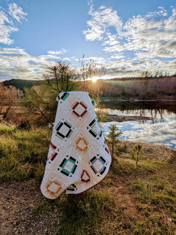 Multi color modern quilt with white background outside at sunset with river in background.