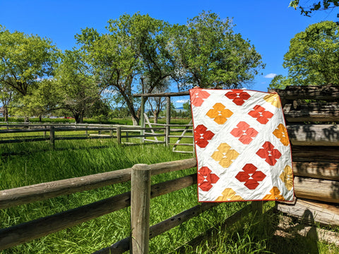 Modern quilt in shades of orange, pink and yellow on fence with trees and blue sky.