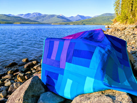 blue and purple quilt on rock next to lake and mountains