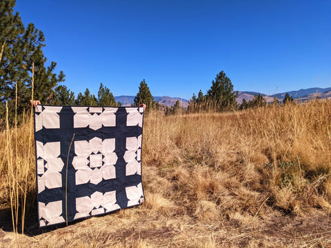 Black and tan large flower quilt in meadow with trees and blue sky.
