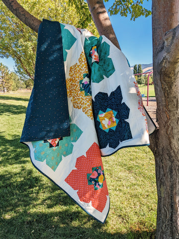 Colorful large retro flower quilt hanging in tree with grass and blue sky.