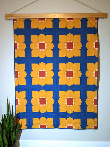 Retro large flower yellow, pink and blue quilt hanging on wall with green plant in front.
