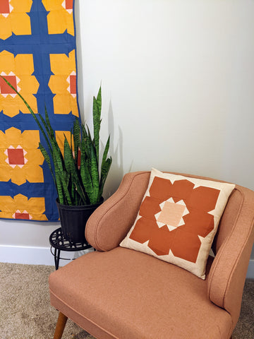 Orange and pink large modern flower pillow on pink chair, infront of yellow and blue flower quilt.