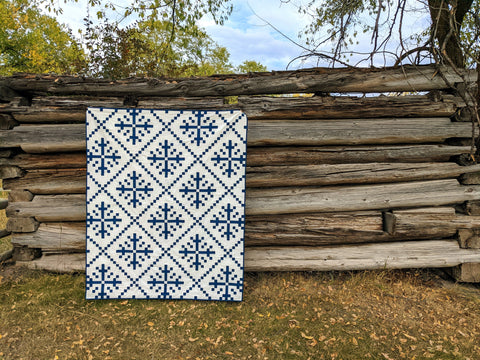 White and blue modern snowflake quilt in front of old log building.