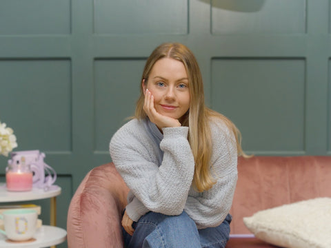 Sophie with blond hair, sitting on a pink couch, with her legs crossed. She is look directly into the camera and is smiling with her head resting on her hand. 