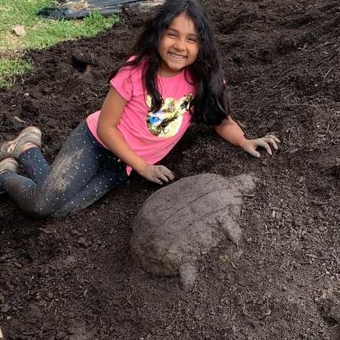 Brown girl with long black hair reclining on a hill of soil near a turtle sculpture she made out of soil.