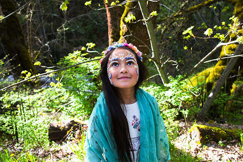 Photoof a young girl in a forest setting, her face adorned with whimsical face painting for kids designs