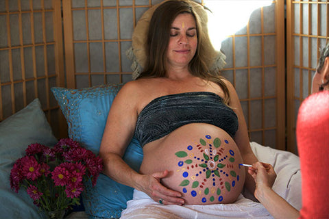 A mother-to-be sitting with her large pregnant belly painted like a radiant sun, an example of creative face painting for kids on an unexpected canvas